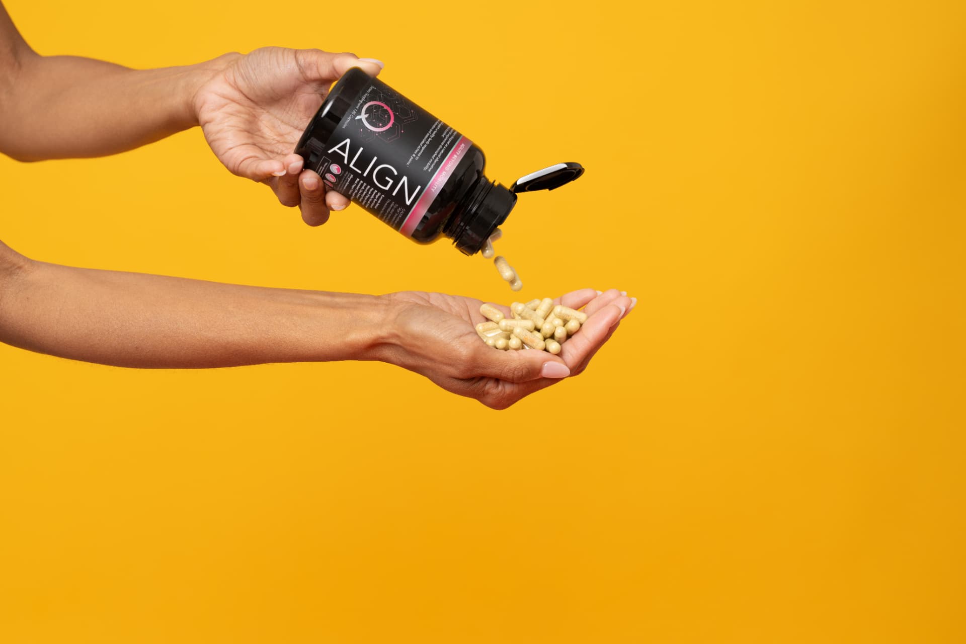 Hands pouring light-colored supplement capsules from a black bottle into a cupped palm against a solid yellow background
