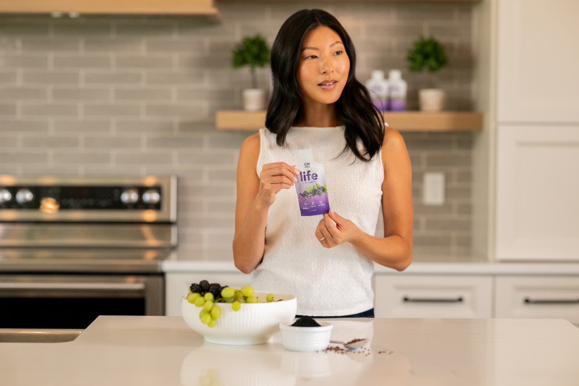 Original Nutrition 'Life' supplement packet held by a woman in a modern kitchen, with a bowl of grapes and blackberries on the counter